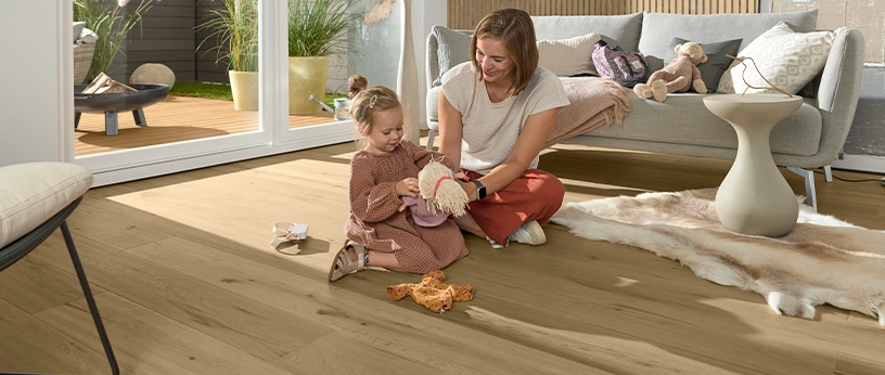 Helles Parkett im Wohnzimmer. Eine Frau spielt mit einem kleinen Mädchen auf dem Boden.