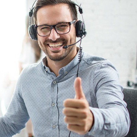 Freundlicher Kundenberater mit Headset sitzt an einem Schreibtisch mit Computer und zeigt den Daumen hoch.