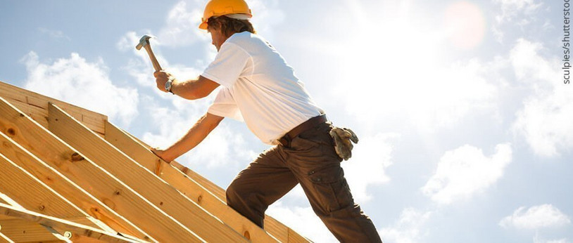 Dachdecker mit gelbem Schutzhelm beim Befestigen von Konstruktionsvollholz (KVH) auf einem Dachstuhl unter strahlendem Himmel.