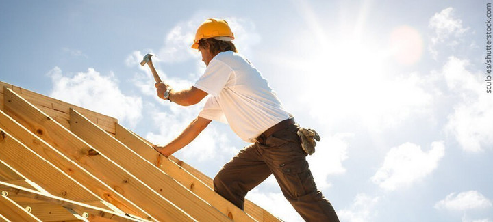 Dachdecker mit gelbem Schutzhelm beim Befestigen von Konstruktionsvollholz (KVH) auf einem Dachstuhl unter strahlendem Himmel.