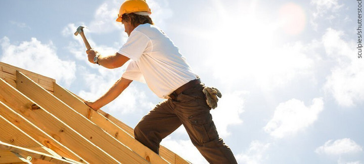 Dachdecker mit gelbem Schutzhelm beim Befestigen von Konstruktionsvollholz (KVH) auf einem Dachstuhl unter strahlendem Himmel.