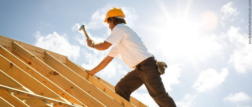Dachdecker mit gelbem Schutzhelm beim Befestigen von Konstruktionsvollholz (KVH) auf einem Dachstuhl unter strahlendem Himmel.