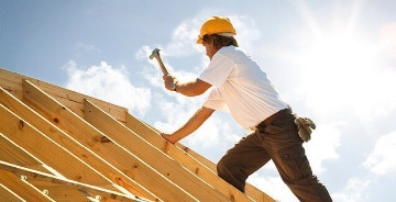 Dachdecker mit gelbem Schutzhelm beim Befestigen von Konstruktionsvollholz (KVH) auf einem Dachstuhl unter strahlendem Himmel.