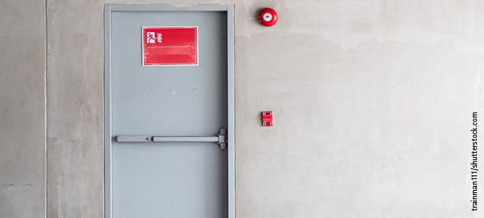 Graue Brandschutztür mit Fluchtwegsymbol und roter Alarmglocke.