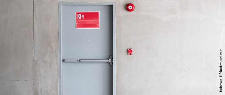 Graue Brandschutztür mit Fluchtwegsymbol und roter Alarmglocke.