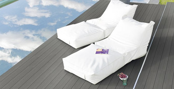 Elegante Poolterrasse mit dunklem WPC-Belag, zwei weißen Lounge-Liegen und ruhiger Wasserreflexion.